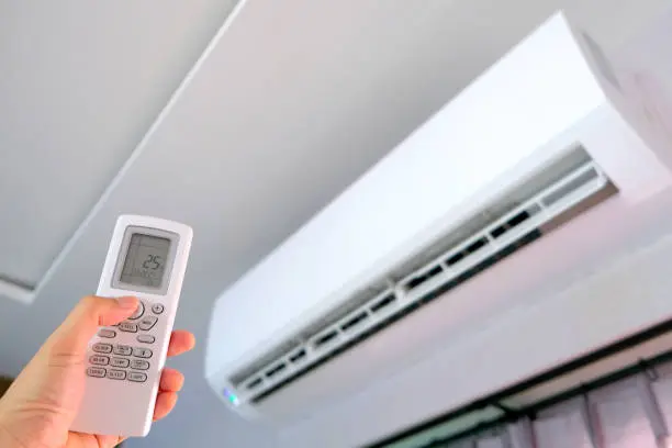 Mano sosteniendo mando a distancia apuntando a split de aire acondicionado en pared con display mostrando temperatura